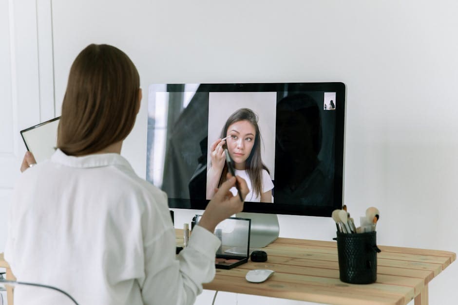 Woman following an online beauty tutorial, applying makeup using brushes in front of a computer screen.