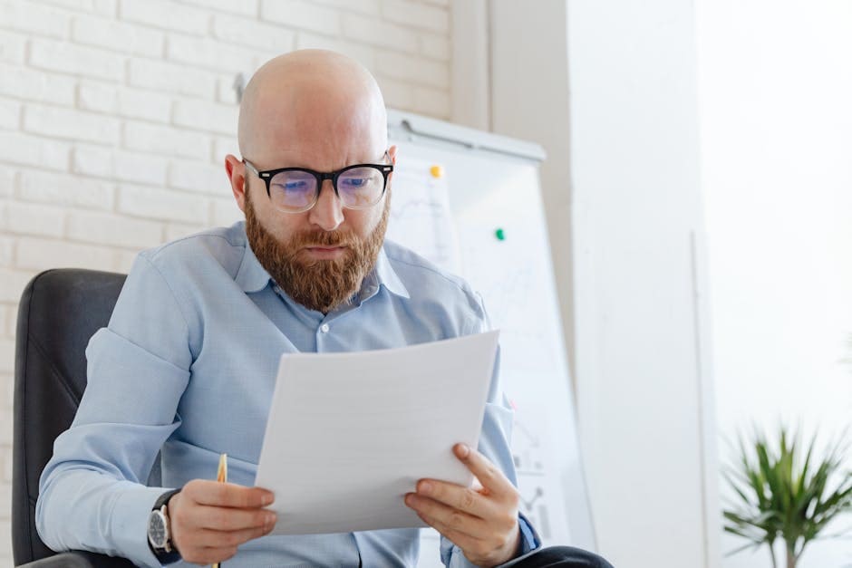 A businessman with a beard and eyeglasses reviewing documents in an office setting.