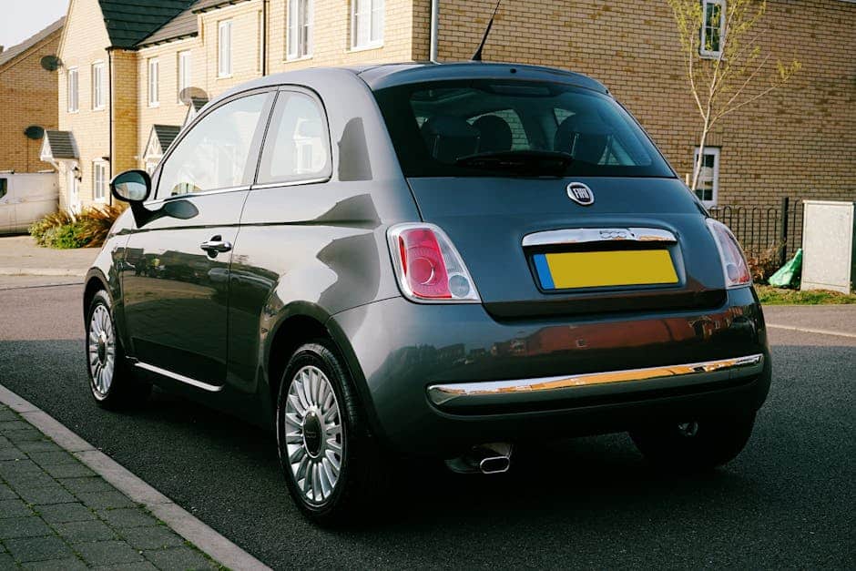 A classic Fiat 500 parked on a suburban street in the UK, featuring a clear sunny day and modern housing.